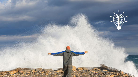 A person standing with arms outstretched facing a large wave crashing on a rocky shore, symbolizing resilience and strength in the face of challenges.